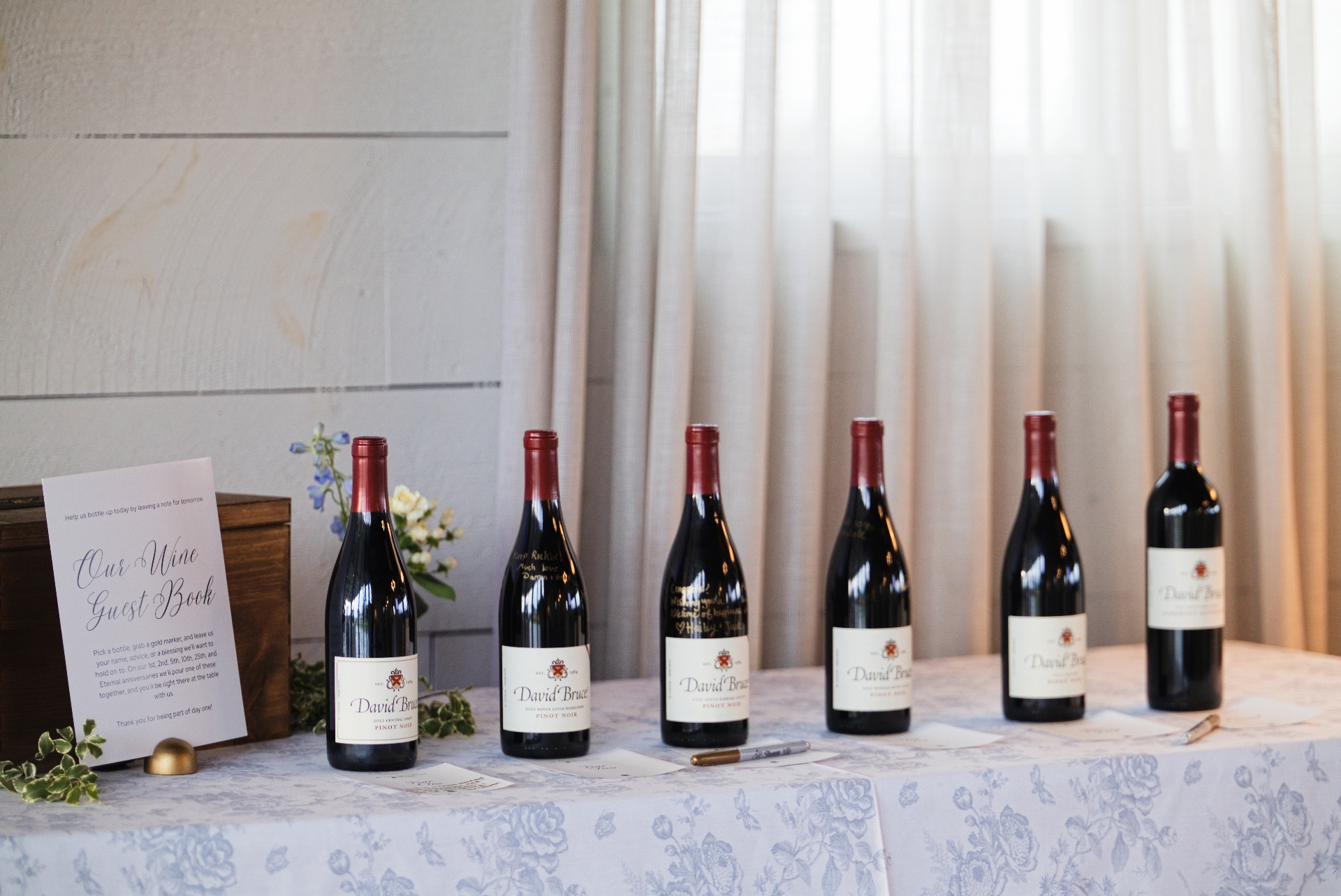 Wedding guest book table with six David Bruce wine bottles, signing cards, and gold pens